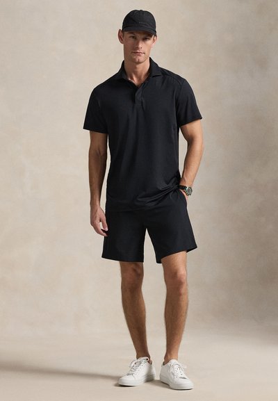 Μαύρο πόλο πουκάμισο και σορτς από αεριζόμενο ύφασμα, συνδυασμένα με λευκά αθλητικά παπούτσια. Το άτομο φοράει μαύρο καπέλο. Ουδέτερο φόντο.