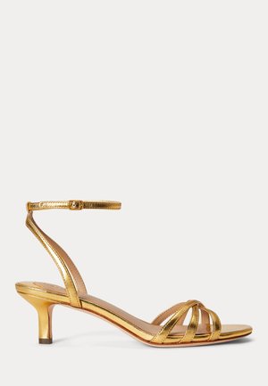 Zlatá metalická sandála s remienkami, nízkym opätkom a nastaviteľnou prackou na členku na čistom bielom pozadí.