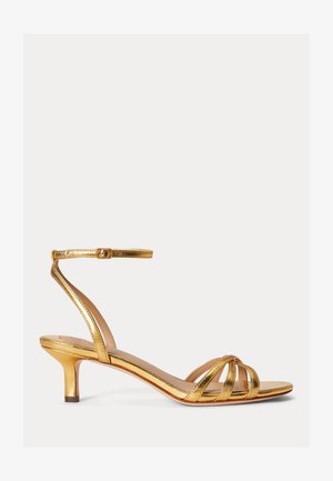 Kuldse metallilise viimistlusega sandaal madala kontsaga, millel on reguleeritava paelaga pahkluu pandla ja valgel taustal.