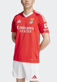 Červený športový dres s výstrihom do V, s textúrovaným zigzagovým vzorom, bielym brandingom a tromi zlatými hviezdami. Skombinovaný s bielymi šortkami.