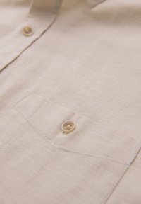 Section de chemise en lin beige montrant une couture détaillée, deux boutons beiges et une poche sur un tissu texturé.