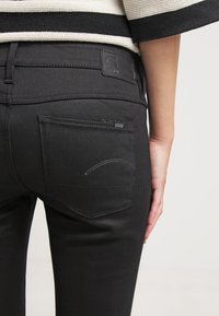 Jeans de mezclilla negra con un corte ajustado, que incluyen un bolsillo trasero y un pequeño parche con el logo en la cintura. La tela tiene una textura suave.