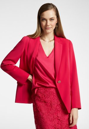 Heldere rode blazer met een gladde textuur en een enkele gouden knoop, gecombineerd met een bijpassende satijnen top en een kanten kokerrok met bloemenpatronen.