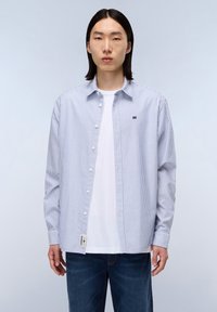 Chemise à rayures bleu clair et blanche, en tissu coton, à manches longues, avec un petit logo sur la poitrine et une étiquette à l'ourlet.