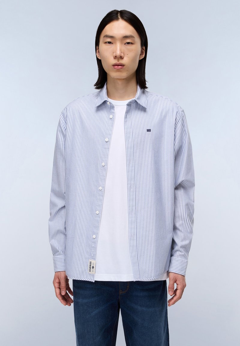 Chemise à rayures bleu clair et blanche, en tissu coton, à manches longues, avec un petit logo sur la poitrine et une étiquette à l'ourlet.