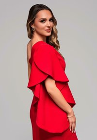 La robe rouge présente des manches superposées et flottantes ainsi qu'une silhouette cintrée. Le tissu a une texture lisse, rehaussée par des accents de bijoux subtils.