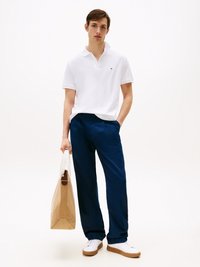 Witte polo met een klein logo, gecombineerd met marineblauwe broeken. Het model houdt een tan tote bag vast en draagt witte sneakers met beige zolen.