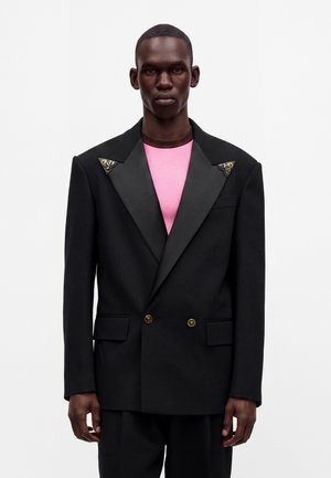 Man draagt een zwarte dubbelgeknoopte blazer met satijnen revers en decoratieve spelden, over een roze overhemd, staand tegen een effen achtergrond.