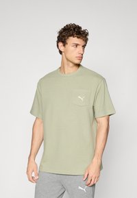 Olijfgroene t-shirt met korte mouwen in geribbelde textuur, met een linkse borstzak waarin een wit Puma-logo is geborduurd. Rond de hals en een losse pasvorm.