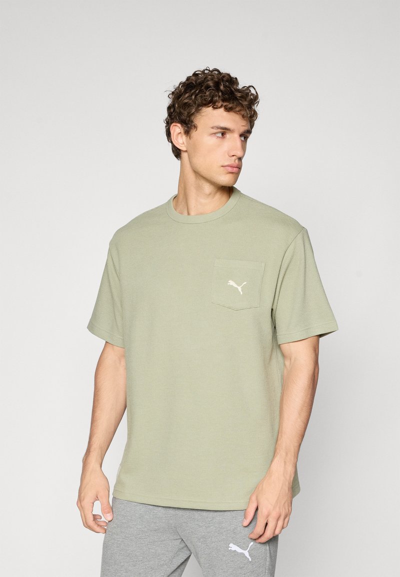 Olijfgroene t-shirt met korte mouwen in geribbelde textuur, met een linkse borstzak waarin een wit Puma-logo is geborduurd. Rond de hals en een losse pasvorm.