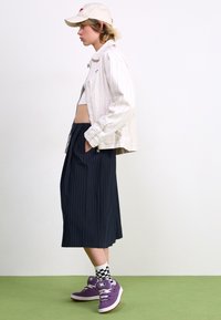 Hvit stripete jakke, cropped topp, mørk stripete skjørt, lilla sneakers og rutete sokker. Plassert mot et grønt gulv og nøytral bakgrunn.