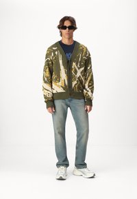 Overdimensjonert grønn cardigan med abstrakte gule og hvite mønstre, båret over en marineblå t-skjorte, kombinert med lyseblå jeans og grå joggesko.