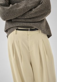 Pull en maille marron rentré dans un pantalon plissé crème taille haute avec une ceinture noire, personne debout les bras croisés.