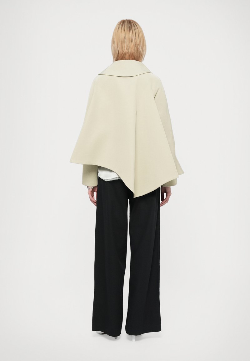 Manteau de style cape en mélange de laine beige clair, avec un large col et une silhouette dramatique. Associé à un pantalon noir à jambes larges.