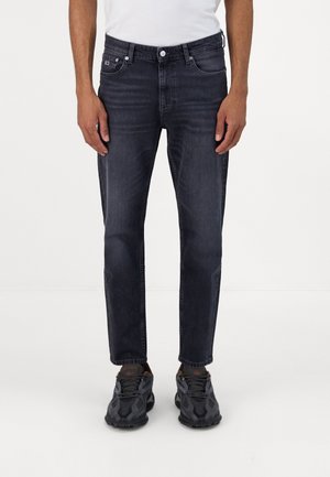 Mężczyzna noszący ciemno prane czarne jeansy slim fit i czarne buty sportowe, stojący na jednolitym białym tle.