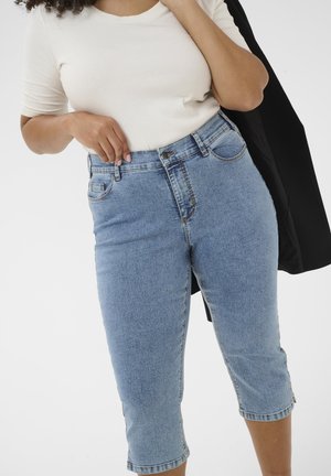 Woman wearing light blue cropped jeans and white short-sleeve top, holding a black jacket over one shoulder against a plain background.