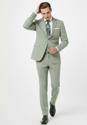 Hombre con traje verde claro ajustándose el botón de la chaqueta, vistiendo camisa blanca, corbata verde con estampado y zapatos de vestir negros, de pie frente a un fondo blanco.