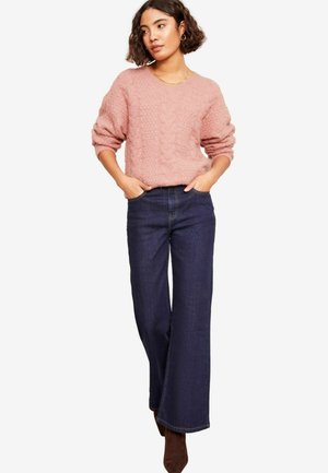 Roze kabeltrui met V-hals, gecombineerd met donkerblauwe wijde spijkerbroek. De outfit omvat bruine enkellaarsjes en gouden creolen.