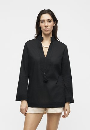 Kobieta w czarnej tunice z długim rękawem, z dekoltem w kształcie litery V ozdobionym frędzlami, oraz kremowych teksturowanych szortach, stojąca na tle jednolitego jasnego koloru.