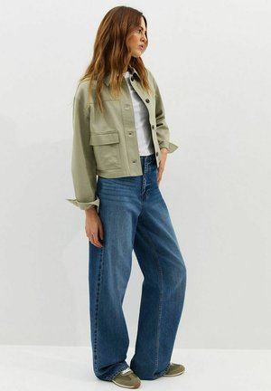 Kobieta w zielonej przygaszonej kurtce typu cropped, białej koszulce, szerokich niebieskich dżinsach i zielonych trampkach stoi na białym, jednolitym tle.