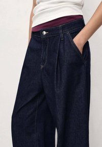 Pantalons en denim bleu foncé à taille haute, avec des plis à l'avant et des poches latérales ; finition avec des coutures contrastantes et un bouton en métal.