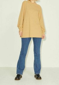 Camicia beige a maniche lunghe con scollatura rotonda abbinata a jeans a zampa d'elefante blu e scarpe nere. Il tessuto sembra morbido con una texture liscia.