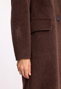 Brązowy wełniany płaszcz o miękkiej, teksturowanej powierzchni. Posiada kieszeń naszywaną oraz jeden czarny guzik. Widoczna dłoń spoczywająca na tkaninie.