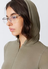 Olive green zip-up hoodie with a fitted design and a soft texture. Features a hood and visible stitching details. Glasses on the model.