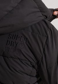 Czarna kurtka puchowa z pikowanym wzorem i tłoczonym logo „SUPER DRY”. Miękki, fakturowany materiał o gładkim wykończeniu i kapturem.