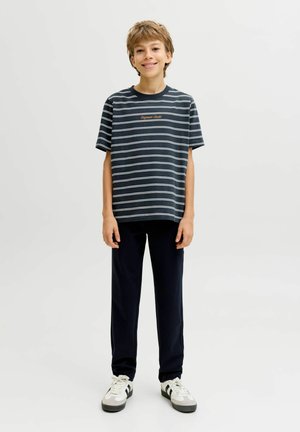 Nasmejan deček stoji oblečen v mornarsko modro in belo črtasto majico, črne hlače in bele športne copate na enostavni svetli podlagi.