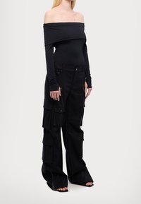 Kvinna iklädd en svart off-shoulder långärmad topp, lösa svarta cargobyxor med flera fickor och svarta öppna klackskor.