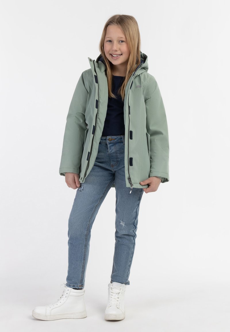 Ljusgrön vattentät jacka med huva, svarta detaljer och dragkedja fram, i kombination med blå jeans och vita högsneakers.