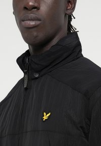 Black lightweight jacket featuring a high collar, textured fabric, and a yellow embroidered logo on the left side. Zippered closure detail.