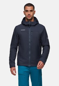 Uomo che indossa una giacca a cappuccio blu navy con zip frontale e pantaloni turchesi, in piedi di fronte a uno sfondo semplice.