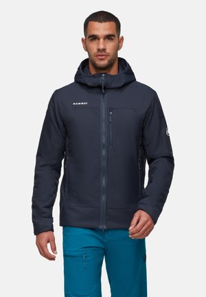 Ein Mann trägt eine marineblaue Kapuzenjacke mit Reißverschluss und tealfarbene Hose, während er vor einem schlichten Hintergrund steht.