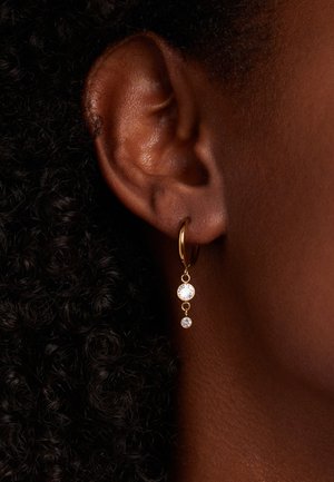 Gold hoop earrings feature two dangling clear crystals in varying sizes, with a smooth finish and minimalist design, worn on a textured ear.