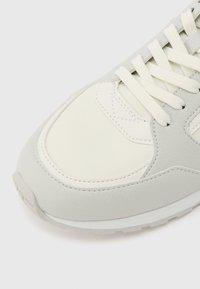 Λευκό και ανοιχτό γκρι μπροστινό μέρος αθλητικού παπουτσιού με υφή από καουτσούκ και λευκά κορδόνια σε απλό ανοιχτό φόντο.