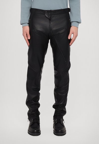 Pantalons en cuir noir ajustés, dotés d'une fermeture éclair, de sangles réglables et de détails de couture distincts, associés à des bottes noires.