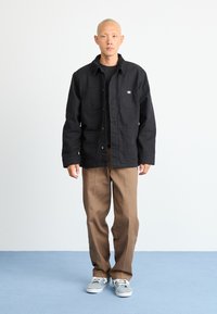 Man standing on blue floor wearing black buttoned jacket, brown pants, and gray sneakers, facing forward against plain white background.