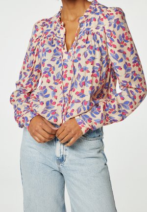 Blouse à manches longues avec motif floral rose et violet, col à volants, et partiellement rentrée dans un jean taille haute bleu clair.
