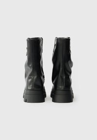 Botas de tobillo de cuero negro con un diseño arrugado, acabado texturizado y suelas de goma negra gruesa. Cuentan con una cremallera lateral para el cierre.