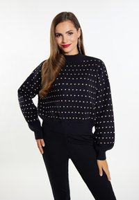 Pulover bleumarin cu un model cu puncte, mâneci lungi și decolteu înalt. Textură tricotată. Purat cu pantaloni negri mulați.