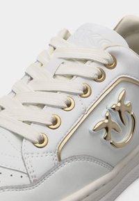 Λευκό αθλητικό παπούτσι με εκρού κορδόνια, χρυσά μεταλλικά τρύπια και λογότυπο πουλιού σε χρυσό και λευκό στην πλαϊνή πλευρά κοντά στα δάχτυλα.