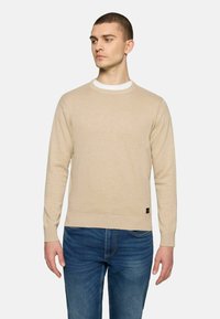 Beige Strickpullover mit klassischem Rundhalsausschnitt, gerippten Bündchen und Saum, kombiniert mit blauen Jeans. Verfügt über ein kleines Markenetikett an der Seite.