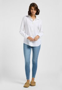 Weißes Hemd mit langen Ärmeln, aus leichtem Stoff, mit Kragen, kombiniert mit hellblauen Skinny Jeans und beigen Hausschuhen.