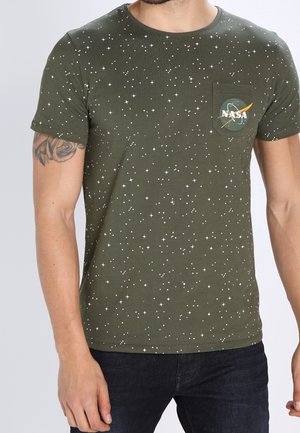 Homme portant un t-shirt vert olive avec un motif d'étoile blanche et le logo NASA sur la poche poitrine, associé à un jean foncé.