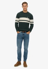 Maglione verde a maglia con strisce bianche, abbinato a jeans blu e stivali marroni con lacci. Posizionato in piedi, mani in tasca.