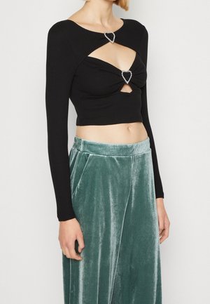 Kobieta ubrana w czarny, długi rękaw crop top z zapięciami w kształcie serca z kryształów oraz zielone aksamitne spodnie o szerokich nogawkach, stojąca na jednolitym tle.