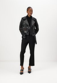 Black leather biker jacket with silver studs and patches, paired with tailored black trousers and black heels featuring buckle accents.