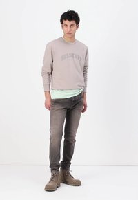 Felpa grigia chiaro con stampa "BELSTAFF", indossata sopra una maglietta verde menta, abbinata a jeans grigi e stivaletti beige.